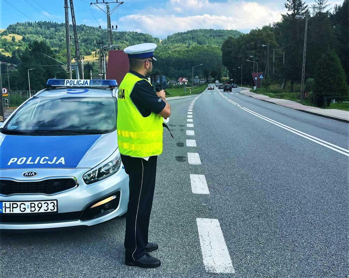 bezpieczeństwo w podróży. umundurowany policjant drogówki z laserowym miernikiem prędkości, obok radiowóz, w oddali jadące samochody